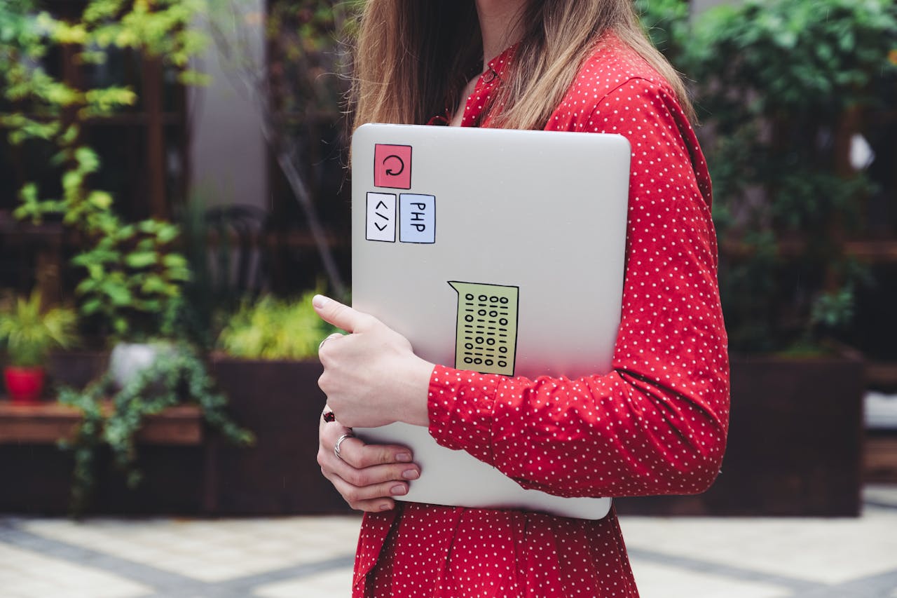 A young woman in a red polka dot shirt holds a laptop with stickers outdoors.